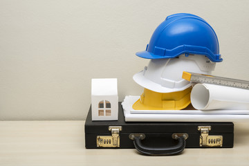 Engineering accessories, helmets, house paper model, business bag, blueprint, on wood table with white concrete wall background. Picture for add text message. Backdrop for design art work.