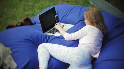 young woman uses a laptop, lying in a lounge area of the park on soft puffs