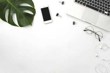 workspace with laptop, mobile phone, green palm branch, glasses, white headphones and clips on white
