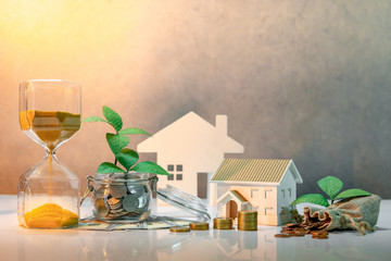 Real estate investment or property ladder. Home mortgage loan rate. Saving money concept. Plant growing out of coins in glass jar with dollar banknotes, hourglass, money bag and house model on table