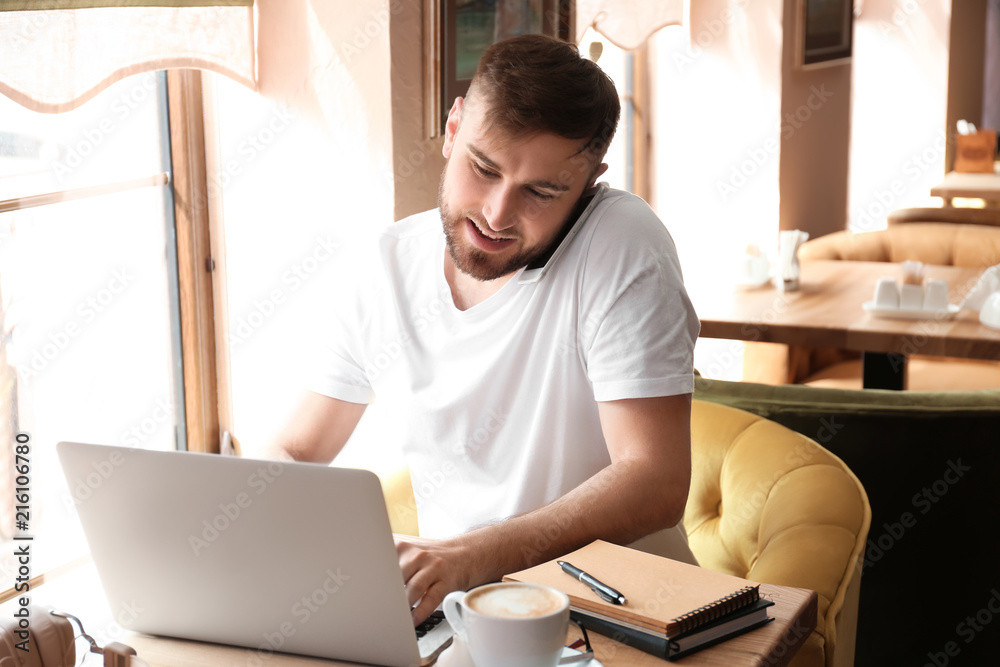 Young freelancer talking on mobile phone while working on laptop in cafe
