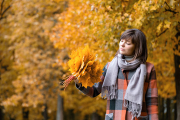 Portrait of young fashion woman outdoor. Brunette woman in autumn park with fashionable plaid coat and scarf. holding yellow leafs