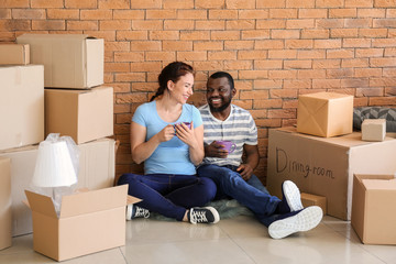 Interracial couple with cups of tea resting on floor near boxes in room. Moving into new house