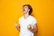 Photo of happy man 20s in casual t-shirt rejoicing and screaming with clenching fists, isolated over yellow background
