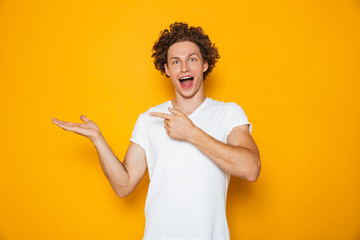 Wall Mural - Young happy man 20s with brown curly hair gesturing finger aside on copyspace at palm, isolated over yellow background