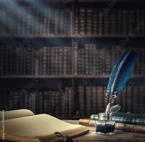 Fotografía Old quill pen, books and vintage inkwell on wooden desk in the old office against the background of the bookcase and the rays of light