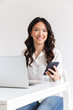 © Drobot Dean - Photo of attractive asian office woman with long dark hair sitting at table and holding smartphone while working with laptop, isolated over white background in studio