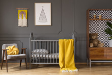 Real Photo Of A Baby Cot With Yellow Cot Standing Between An Armchair And A Cupboard With Teddy Bear And A Plant In Child's Room Interior