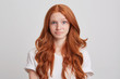 Portrait of confused astonished young woman with long wavy red hair and freckles wears t shirt feels embarrassed and looks directly in camera isolated over white background