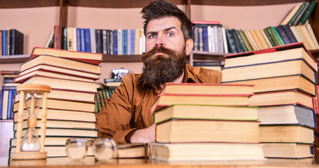 Leinwandbilder - Man on strict face sit between piles of books, while studying in library, bookshelves on background. Teacher or student with beard sit at table with hourglass and glasses, defocused. Bookworm concept.