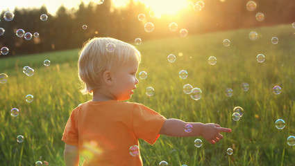 Cute toddler blond boy playing with soap bubbles on summer field. Beautiful sunset light. Happy childhood concept. Authentic lifestyle image