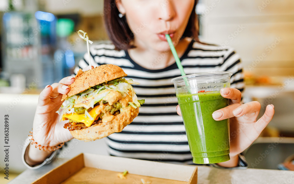 A young woman drinks green smoothies and eats a burger in a vegan fast food restaurant - obrazy, fototapety, plakaty 