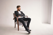 Indoor Portrait Of Handsome Young Male Lawyer With Thick Beard And Trendy Hairstyle Sitting Comfortably In Armchair, Keeping Arms Crossed On Chest And Looking Away With Pensive Thoughtful Expression
