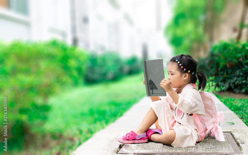 little girl reading a book with backpack sitting in the park ready back ...
