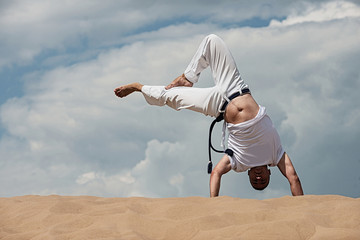 A young guy trains capoeira against the sky. A man performs an acrobatic trick