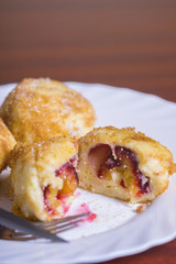 Fresh plum dumplings. One sliced dumpling and two whole with knife and fork on white plate on dark red table