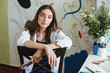 Pensive girl in white shirt on chair thoughtfully looking in camera while holding paint brushes in hand with big painting on background at home