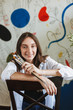 Cheerful girl in white shirt on chair joyfully looking in camera while holding paint brushes in hand with big patterns painting on background at home