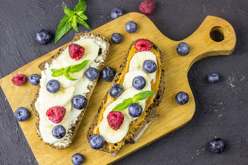 Wall Mural - Traditional American and European summer breakfast: sandwiches of toast with peanut butter or cream cheese with raspberries and blueberry, banana. Black concrete table. copy space top view