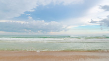 A Front View Of A Beach With The Sand And A Green Sea With Small Waves With A Blue Sky With Some Clouds In The Background