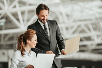 Poster - Smiling unshaven boss telling with outgoing worker while pointing at document in folder. Happy lady looking at it indoor