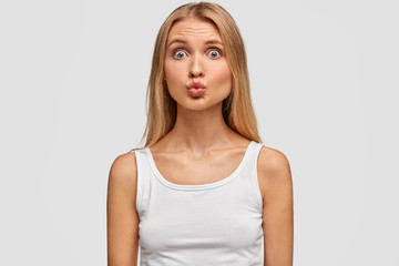 Adorable blonde woman with healthy skin and eyes popped out, keeps lips round, makes grimace, dressed in casual white t shirt, has slim figure, poses over studio blank wall. Facial expressions, beauty