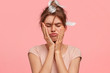 © WHstudio Leushin N - Displeased European female with gloomy expression, purses lips, closes eyes, keeps both hands on cheeks, doesn`t have enough sleep and good rest, stands against pink background. Morning time