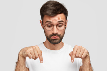 Wall Mural - Headshot of handsome bearded male points down with surprised look, notices something on floor, wears glasses, dressed in casual t shirt, isolated over white background. People and astonishment