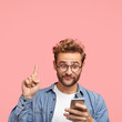 © wayhome.studio  - Vertical shot of curious male has appealing appearance, raises fore finger, looks with great interest at smart phone, reads relevant news in internet, isolated over pink background with copy space
