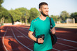 Young man in wireless earphones holding sport bottle in hand happily looking aside on running track of stadium