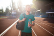Young smiling man joyfully looking aside with sport bag on shoulder and bottle of water in hand on running track of stadium