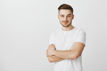 Wall Mural - Intrigued smart young male entrepreneur in white t-shirt with moustache smirking holding hands crossed on chest feeling satisfied and glad receiving great result in sales over gray background