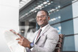 © xartproduction - black man businessman in a business suit and glasses sits on a bench and reads a newspaper against the backdrop of a modern city