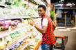 © sofiko14 - African man shopping in vegetable section at supermarket. Black man doing shopping at market while buying vegetables. Handsome guy holding shopping basket reading nutritional values of product.