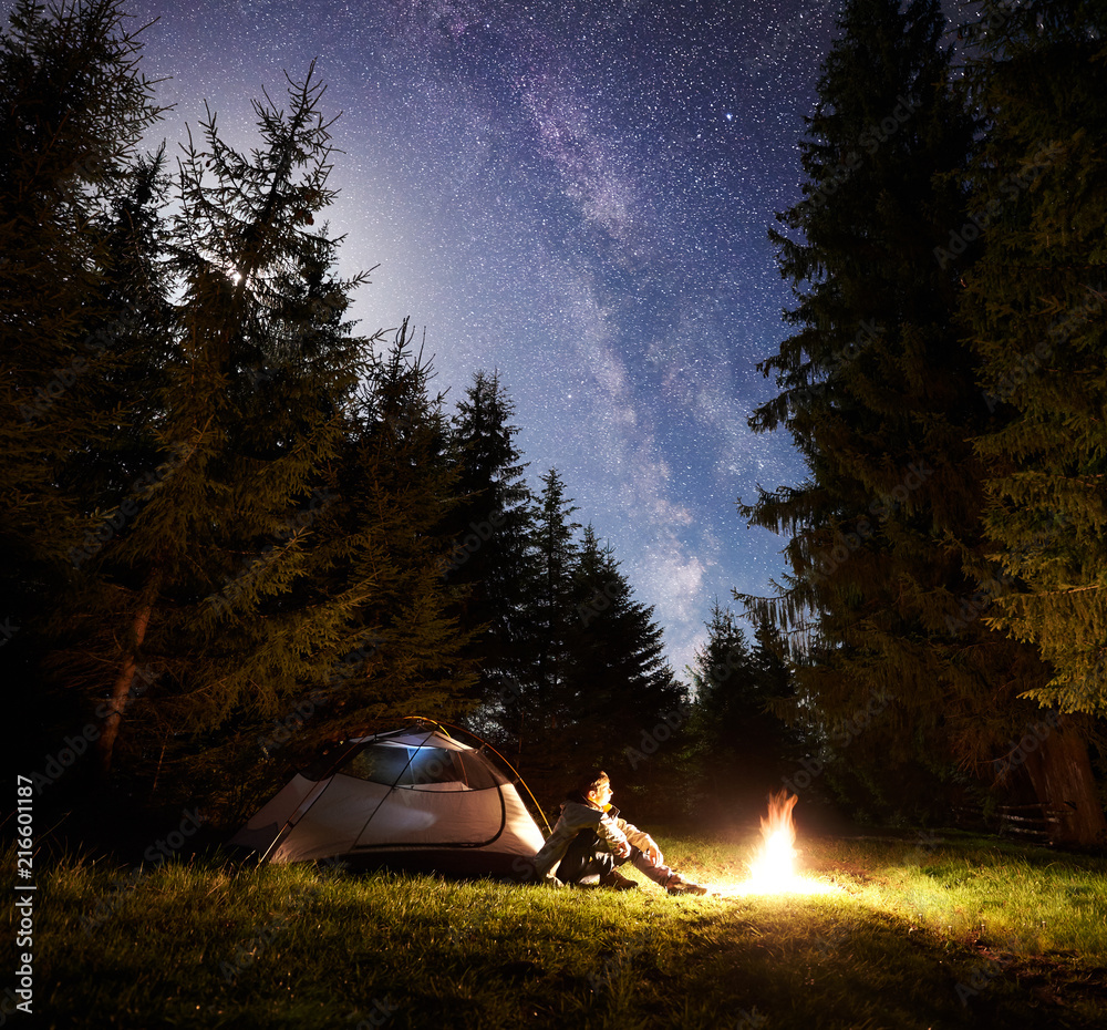 Camping site at night. Tourist tent on forest clearing and male hiker sitting in front of burning bonfire under night blue starry sky on pine trees background. Beauty of nature and tourism concept. - obrazy, fototapety, plakaty 