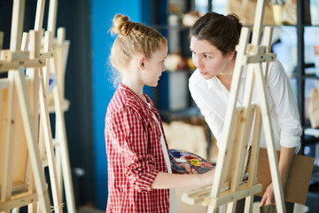 Wall Mural - Little girl with color palette listening to her teacher while both standing by easel at lesson of painting