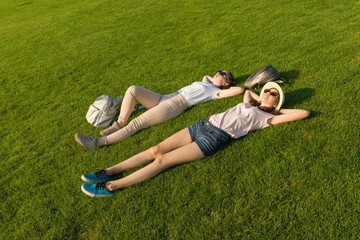 Two young female students with backpacks lying on green lawn grass. Top view, side view.