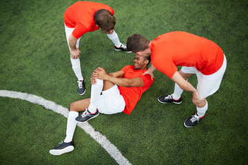 two young football players in uniform leaning over their mate lying on field with hurt knee