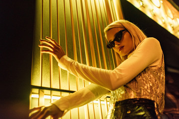 Wall Mural - bottom view of stylish young woman in sunglasses and glossy top over turtleneck on street at night under yellow light