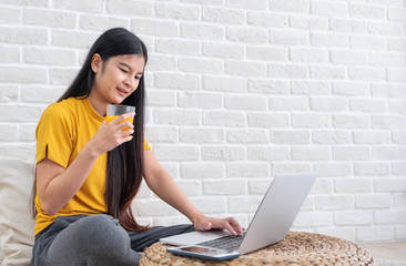Poster - Asian female sit on floor using laptop on wicker stand and drink orange juice in living room at home with relax emotion.Work at home concept.work from home.relax lifestyle.