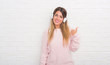 © Krakenimages.com - Young adult woman over white brick wall wearing headphones listening to music and dancing pointing and showing with thumb up to the side with happy face smiling