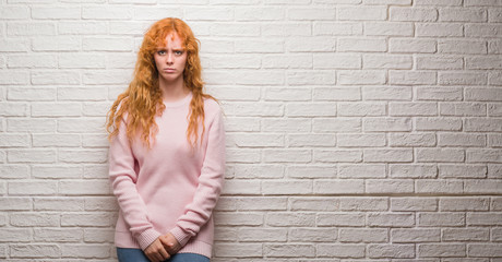 Wall Mural - Young redhead woman standing over brick wall with serious expression on face. Simple and natural looking at the camera.