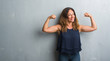 Middle age hispanic woman standing over grey grunge wall showing arms muscles smiling proud. Fitness concept.