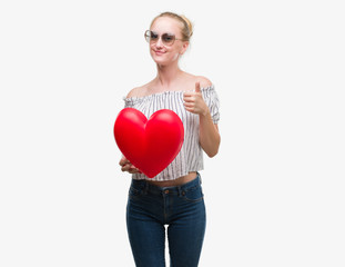 Sticker - Blonde teenager woman holding red heart happy with big smile doing ok sign, thumb up with fingers, excellent sign