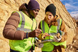 © Seventyfour - Waist up portrait of two industrial workers wearing reflective jackets, one of them African, inspecting mineral ore on site outdoors and using digital tablet