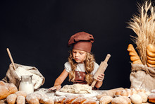 Charming Little Girl With Curly Hair In White Apron And Hat Standing At Table Kneading Bread Dough. KId In Good Mood, Having Fun.