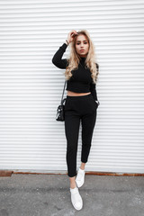 Wall Mural - Young beautiful fashion model girl with a handbag in a black T-shirt, pants and white sneakers poses near a white metal wall