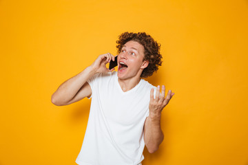 Poster - Photo of happy young guy with curly hair shouting and talking on smartphone, isolated over yellow background