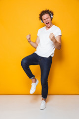 Poster - Full length photo of young smiling guy with curly hair listening to music via headphones and singing, isolated over yellow background