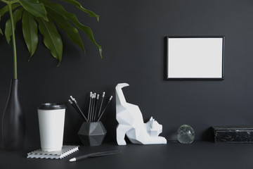 Black design room with mock up poster frame, cat figures, tropical leaf and office accessories. Modern and stylish black interior.